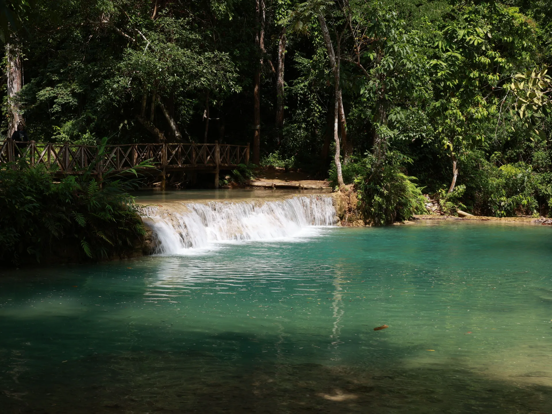 Turquoise pools and a low cascade at Kuang Si Falls, with a timber footbridge under forest canopy